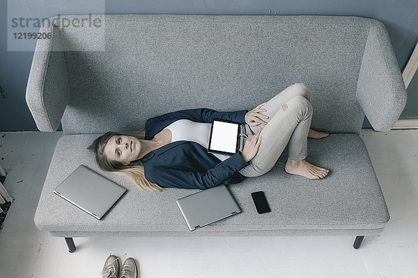 Geschäftsfrau auf der Couch liegend mit mehreren elektronischen Geräten