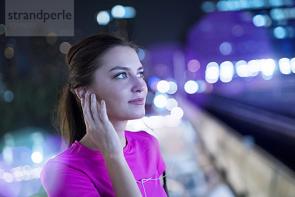 Junge Frau im rosa Sportshirt beim Musikhören in der Stadt bei Nacht
