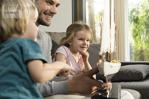 Lächelnder Vater und Kinder beim Betrachten des Spielzeugmodells Schiff auf der Couch zu Hause