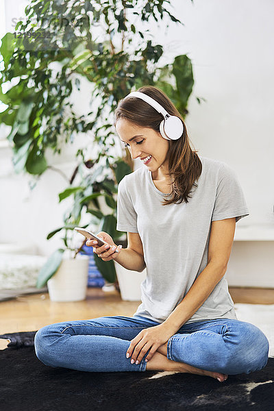 Lächelnde junge Frau auf dem Boden sitzend mit Kopfhörer und Handy