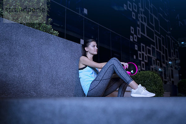 Junge Frau mit rosa Kopfhörern sitzt nachts auf dem Boden in moderner urbaner Umgebung.