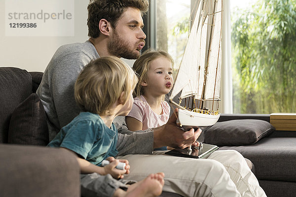 Vater und Kinder blasen in die Segel eines Spielzeugmodellschiffes auf der Couch zu Hause.