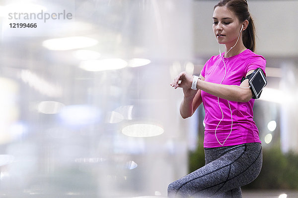 Junge Frau im rosa Sportshirt beim Musik hören und ihre Smartwatch im modernen urbanen Umfeld testen.