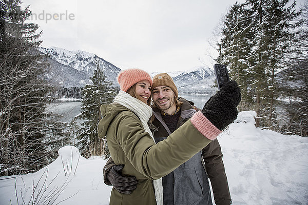 Lächelndes Pärchen bei einem Selfie in alpiner Winterlandschaft mit See