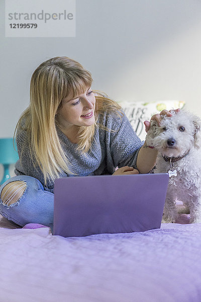 Frau mit Laptop sitzt auf dem Bett und streichelt ihren Hund.