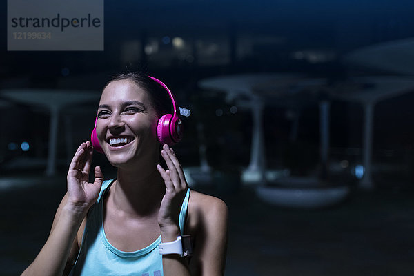 Fröhliche junge Frau mit rosa Kopfhörern  die nachts in der modernen Stadt Musik hört.