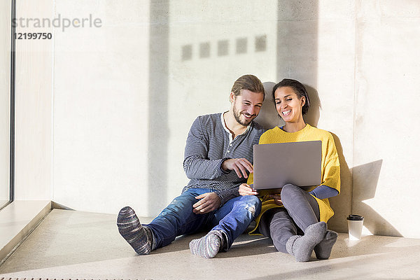 Porträt eines lachenden jungen Paares auf dem Boden sitzend mit Laptop