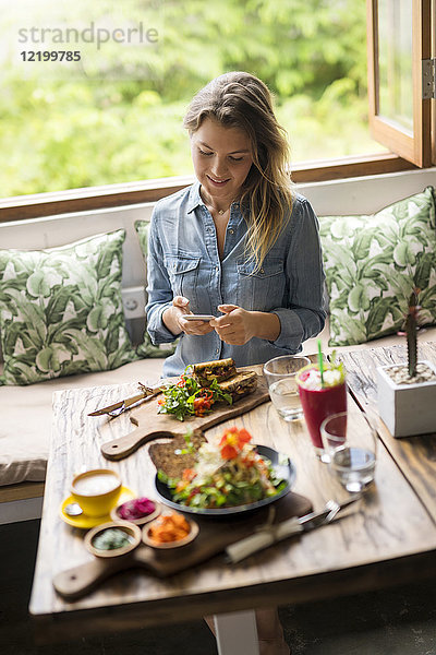 Frau fotografiert Essen mit Smartphone im gemütlichen Café vor dem Fenster