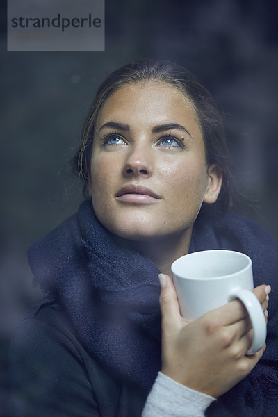 Porträt einer jungen Frau mit Kaffeetasse aus dem Fenster schauend