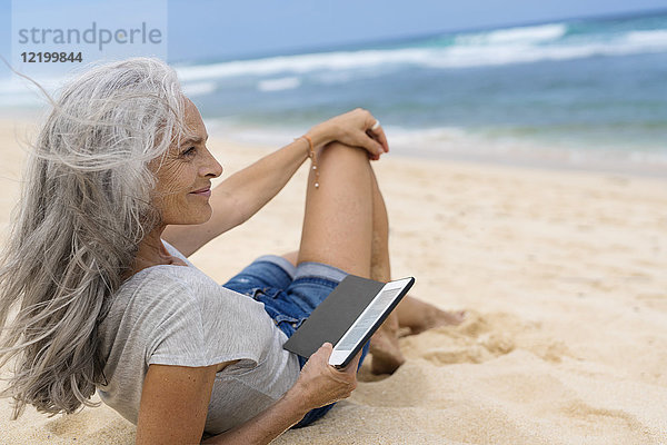 Schöne lächelnde Seniorin am Strand liegend  mit E-Book