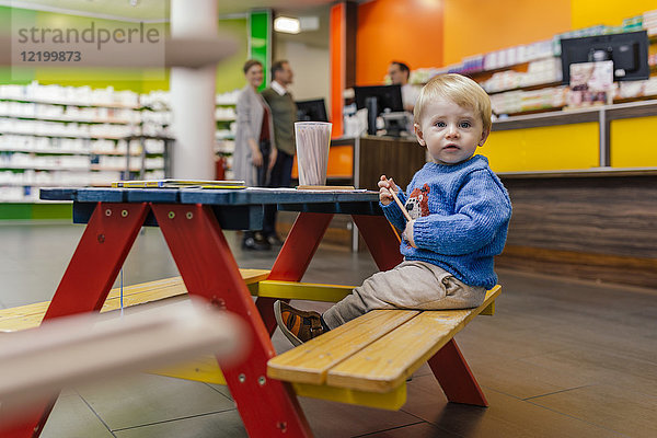 Kleiner Junge auf Bank in der Apotheke mit Eltern im Hintergrund