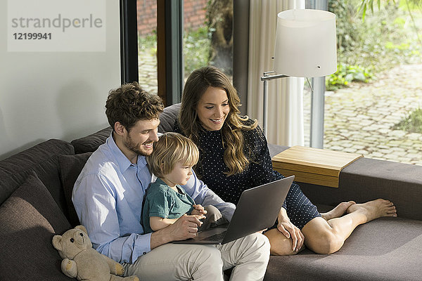 Lächelnde Eltern und Sohn sitzen auf dem Sofa und benutzen den Laptop zu Hause.