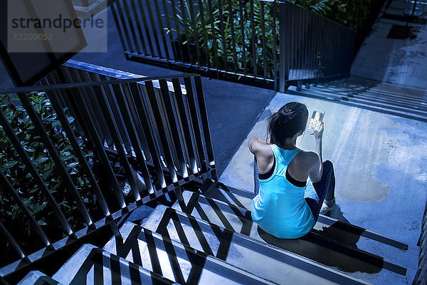 Junge Frau sitzt auf einer Treppe und überprüft nachts ihr Smartphone in einer modernen  urbanen Umgebung.