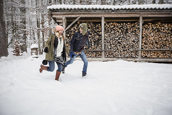Glückliches Paar  das im Winter vor einem Holzhaufen im Freien läuft.