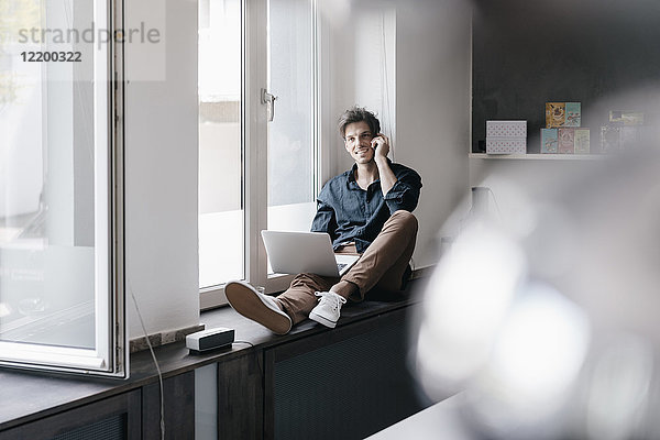 Lächelnder junger Mann am Telefon sitzend mit Laptop auf Fensterbank im Loft