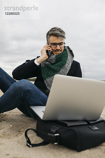Porträt eines Geschäftsmannes am Telefon  der im Winter am Strand sitzt und einen Laptop benutzt.