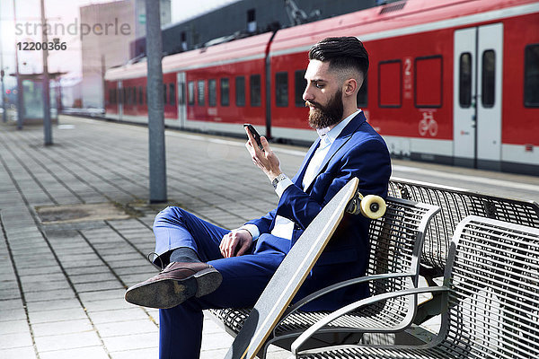 Junger Mann mit Longboard am Bahnhof sitzend  mit Smartphone