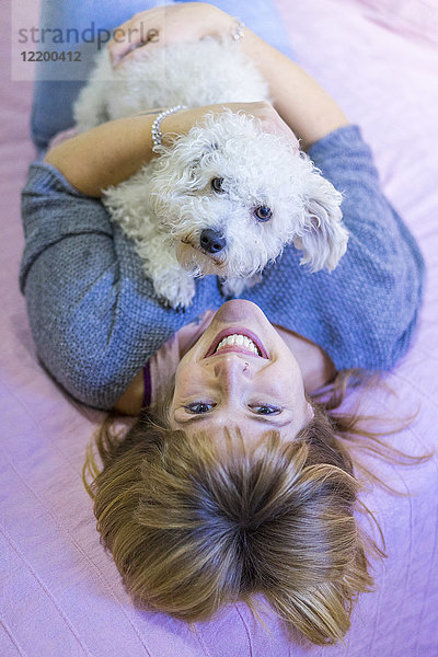 Porträt einer glücklichen Frau  die mit ihrem Hund auf dem Bett liegt.