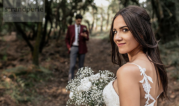 Porträt der lächelnden Braut mit Blumenstrauß im Wald mit Bräutigam im Hintergrund