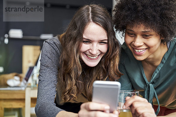 Zwei lächelnde junge Frauen teilen sich ihr Handy im Büro.