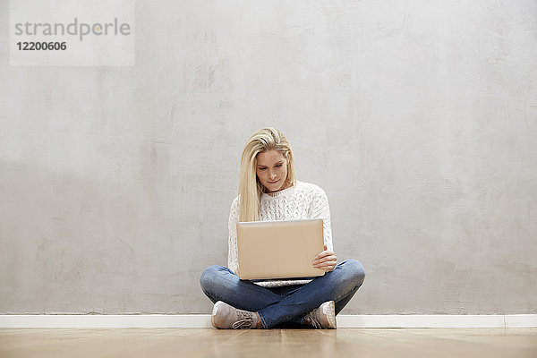 Blonde Frau auf dem Boden vor grauer Wand sitzend mit Laptop