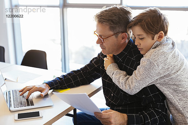 Geschäftsmann mit Laptop am Schreibtisch im Büro mit einem Sohn  der ihn umarmt.