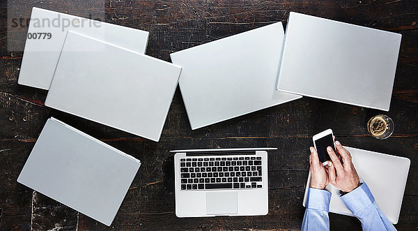 Männerhände mit Smartphone auf Tisch mit sieben Laptops  Draufsicht