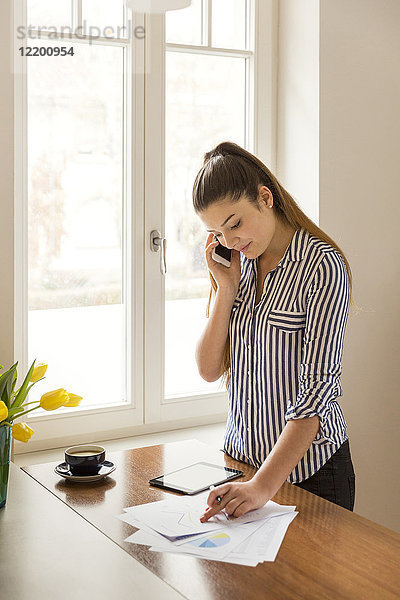Junge Frau am Telefon bei der Arbeit an gedruckten Karten auf einem Tresen zu Hause