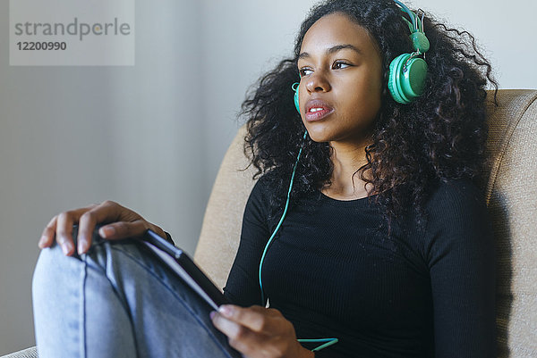 Portrait einer jungen Frau  die im Sessel sitzt und Musik mit Kopfhörer und Tablett hört.