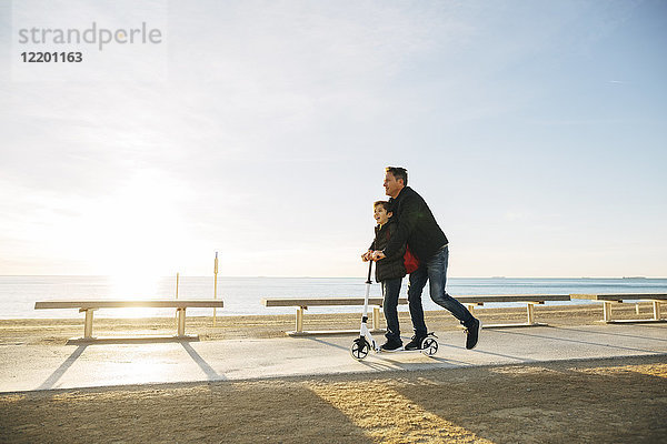 Vater und Sohn fahren Roller auf der Strandpromenade bei Sonnenuntergang
