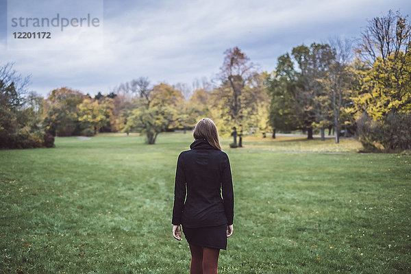 Rückansicht einer jungen Frau  die auf einer Wiese im Herbstpark spazieren geht.