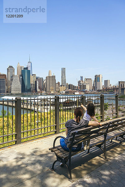 USA  New York  Brooklyn  Rückansicht der Frau und des jungen Mädchens  die auf der Bank sitzen  während sie die Skyline von Manhattan von Brooklyn aus betrachten.
