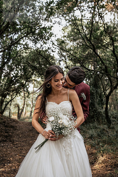 Bräutigam küsst lächelnde Braut im Wald