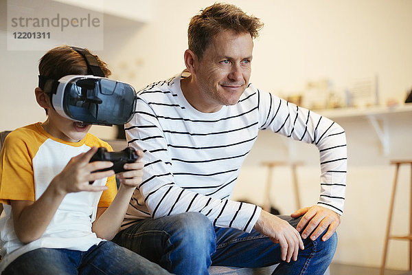 Junge mit VR-Brille beim Videospiel mit Vater auf der Couch zu Hause