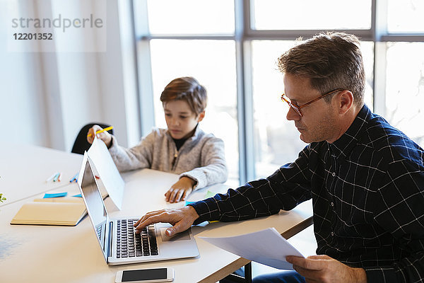 Geschäftsmann mit Laptop am Schreibtisch im Büro  neben ihm sitzt der Sohn