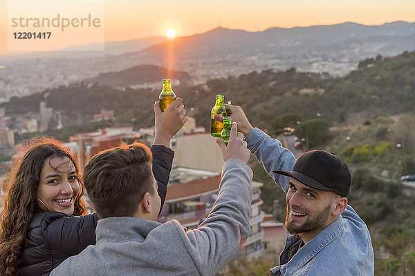 Spanien  Barcelona  drei glückliche Freunde mit Bierflaschen auf einem Hügel mit Blick auf die Stadt bei Sonnenuntergang