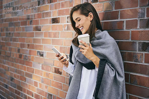 Fröhliche Frau mit Kaffee  um vor der Ziegelwand auf das Handy zu schauen.