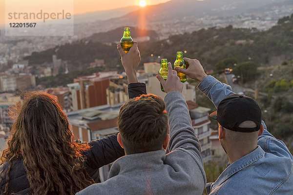 Spanien  Barcelona  drei Freunde mit Bierflaschen auf einem Hügel mit Blick auf die Stadt bei Sonnenuntergang