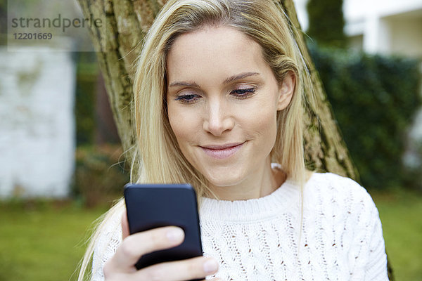 Porträt einer lächelnden blonden Frau beim Blick auf das Handy im Freien