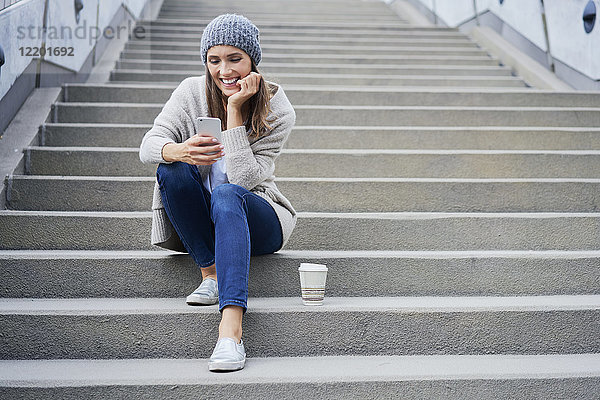 Fröhliche Frau mit Kaffee zum Sitzen auf der Treppe mit Blick auf das Handy