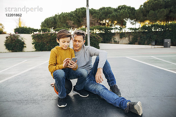 Vater und Sohn sitzen auf dem Basketballfeld mit dem Handy