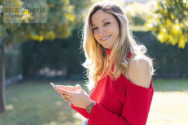 Porträt einer lächelnden jungen Frau mit Smartphone im Garten