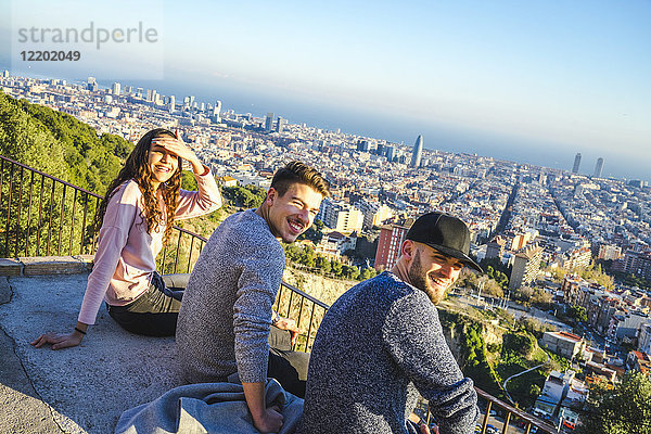 Spanien  Barcelona  drei lächelnde Freunde sitzen auf einer Mauer mit Blick auf die Stadt.