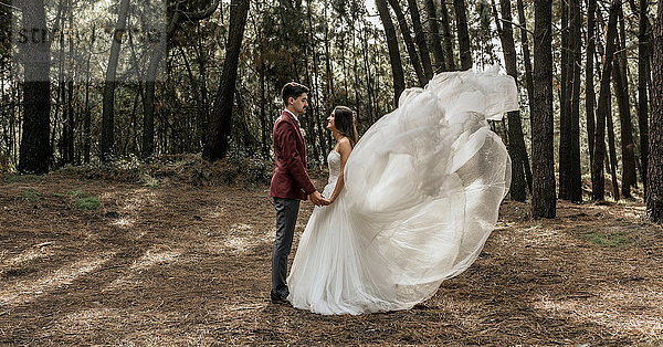 Braut mit windgepeitschtem Brautkleid und Bräutigam im Wald stehend  Händchen haltend