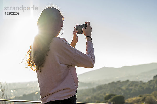 Junge Frau steht auf einem Hügel und macht ein Handyfoto.