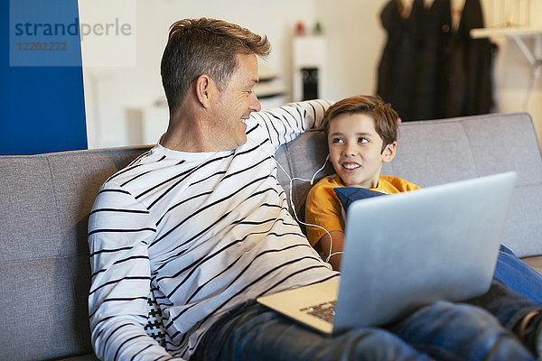 Lächelnder Vater und Sohn mit Ohrstöpseln und Laptop auf der Couch zu Hause