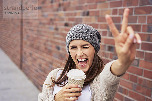 Portrait einer jungen Frau mit Kaffee zum Mitnehmen mit Siegeszeichen