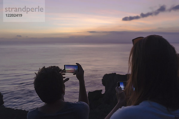 Indonesien  Bali  Lembonganische Insel  Freunde an der Meeresküste in der Abenddämmerung beim Fotografieren mit dem Handy