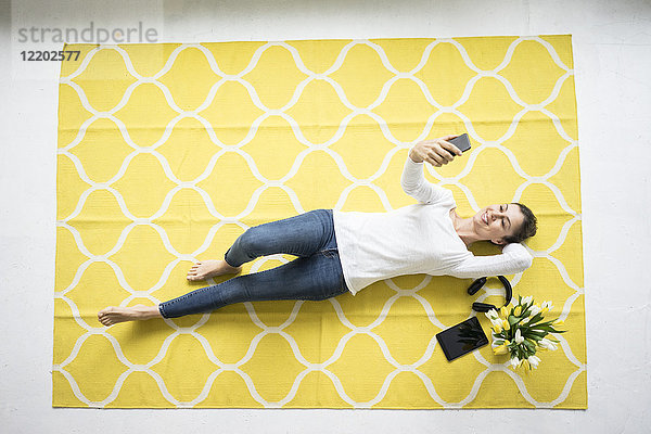 Lächelnde Frau hält Handy auf Teppich liegend mit Tulpenstrauß