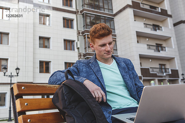 Rothaariger junger Mann auf der Bank sitzend mit Laptop